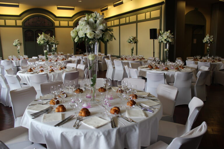 Salle de réception avec tables rondes dressées, nappes blanches, housses de chaises et verrerie – RSB Location